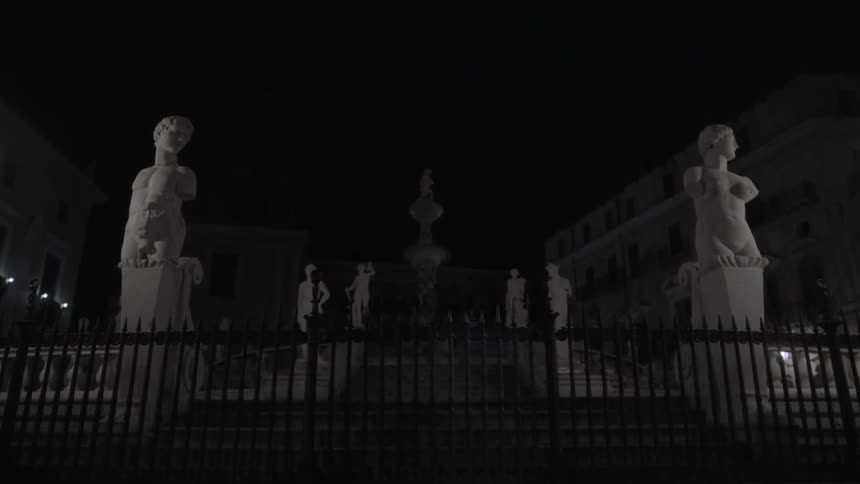 An elaborate marble fountain with statues and an iron fence is seen at night