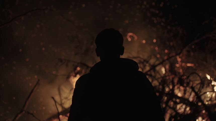 A man wearing a hooded jacket stands staring at a burning fire during the night
