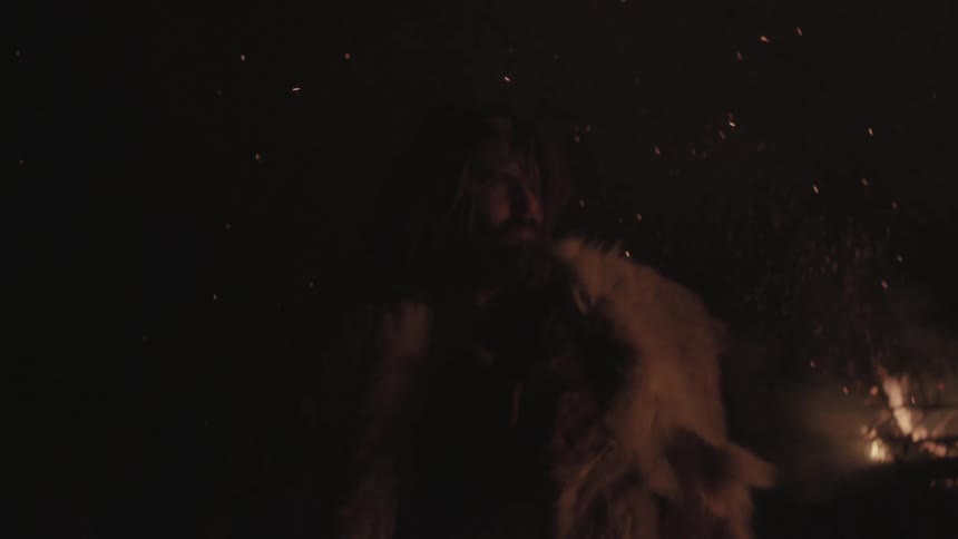 A man, wearing a heavy coat, is standing next to a fire as the embers fall around him