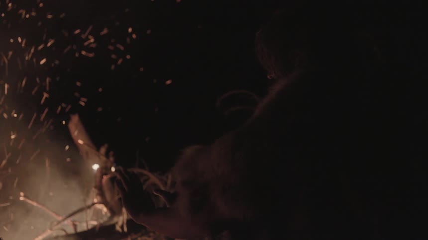 Person warming their hands near a brush fire as sparks float in the air
