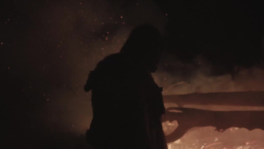 A man stands in front of a raging fire at night