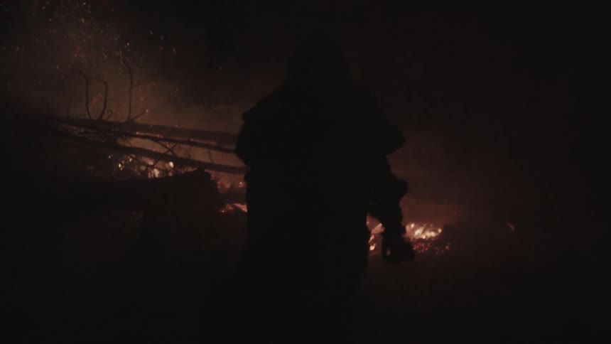It is night and a man wearing furs is approaching a pile of trees and branches that is burning