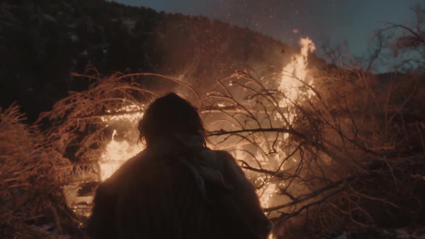 A man stands before a large pile of burning tree branches