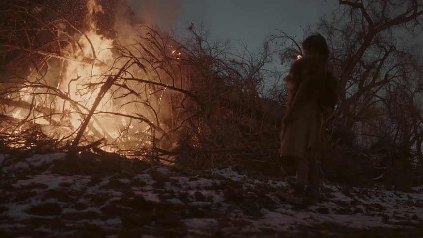 A man stands before a large pile of burning tree branches at night