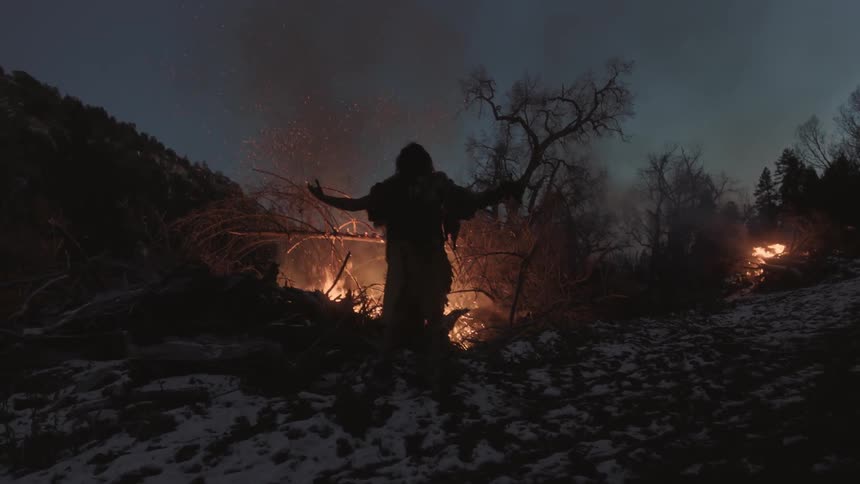 A man stands by a fire before lifting his arms overhead
