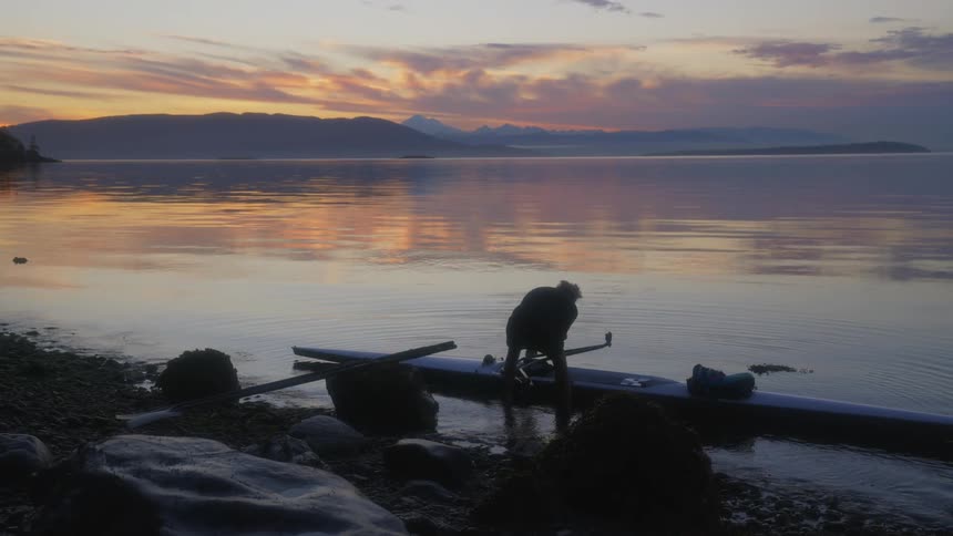 Man sets kayak into water reflecting the red sky and nearby shore