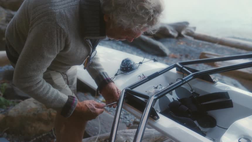 An elderly man stands on the shore and tightens a screw on his kayak