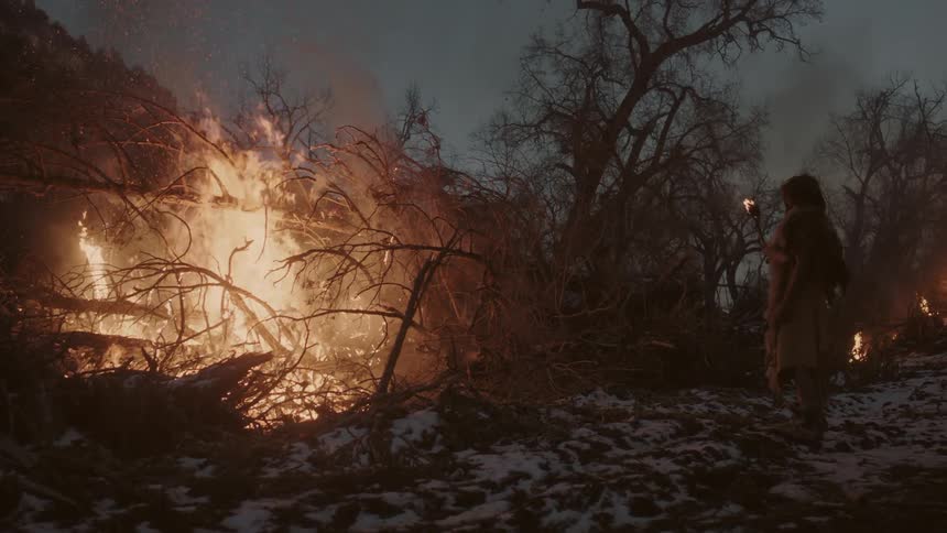 Man stands holding torch watching a large brush pile burn