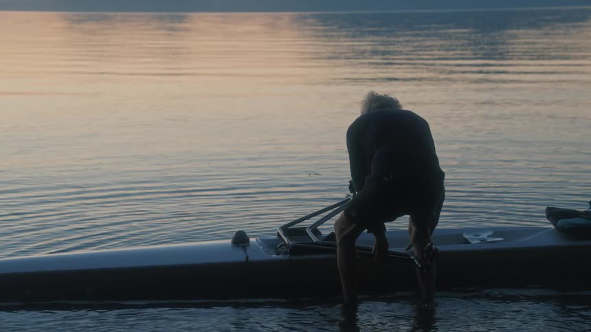 Elderly man lowers a kayak to rest on the surface of a lake at morning