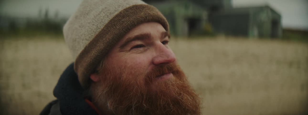 Redheaded man with beard smiles standing in crop field with farmhouse in the background