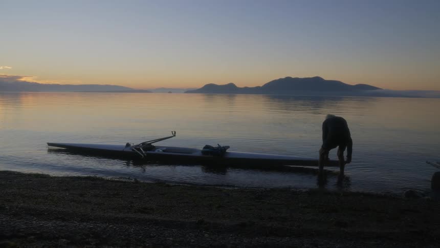 Man places a canoe on surface of a lake next to the coast in front of the mountains at evening