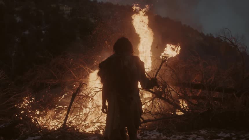 A man carrying a lighted torch stands in front of a large bonfire