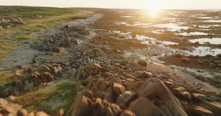 Sun sits on horizon shining across rocky terrain full of puddles and pools bordering green landscape