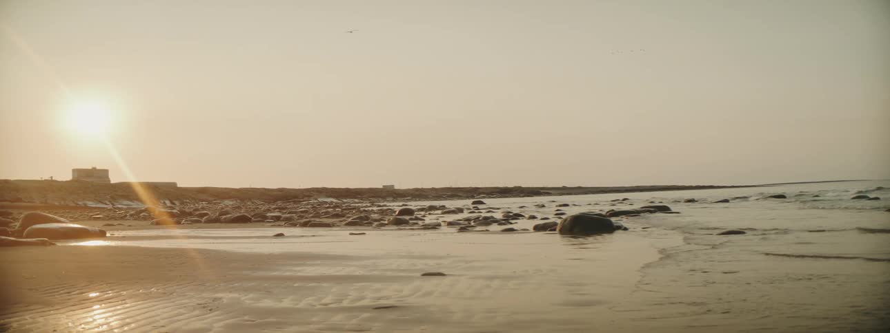 Waves roll into shore on a rock covered beach as the sun shines in a hazy sky as a small boat is on shore