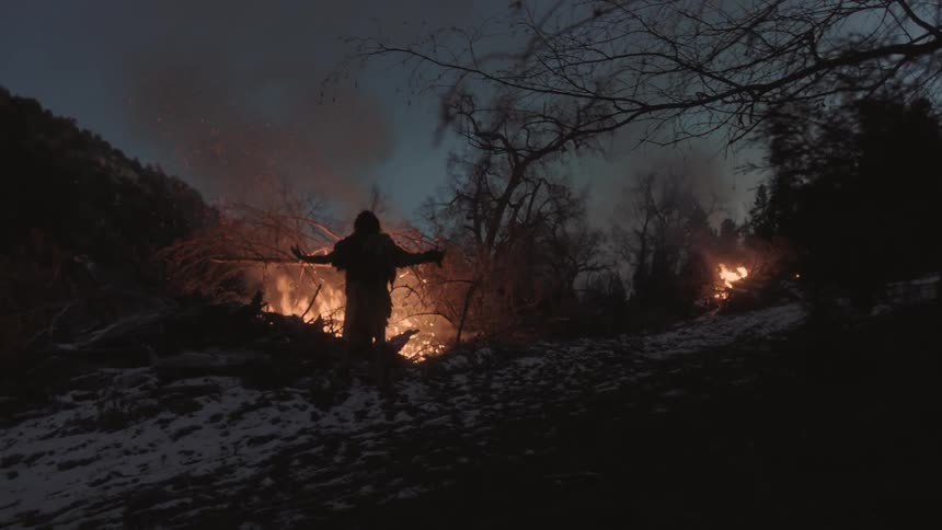 A man stands near a great bonfire and lifts his arm up toward the sky