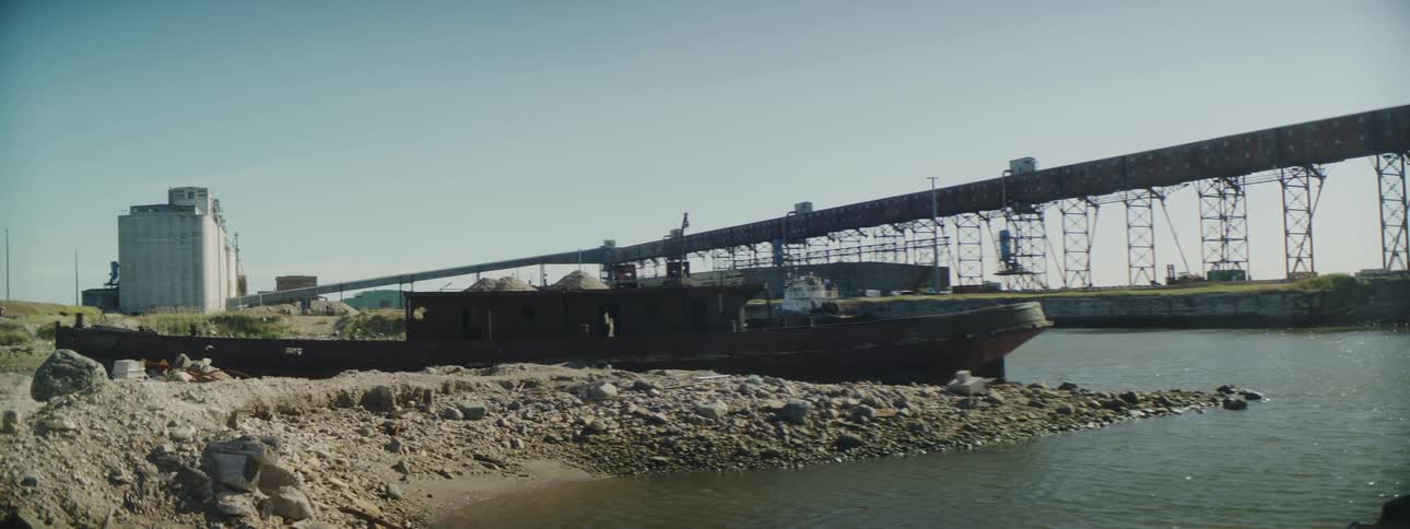 A long boat sits parked along a river near a bridge