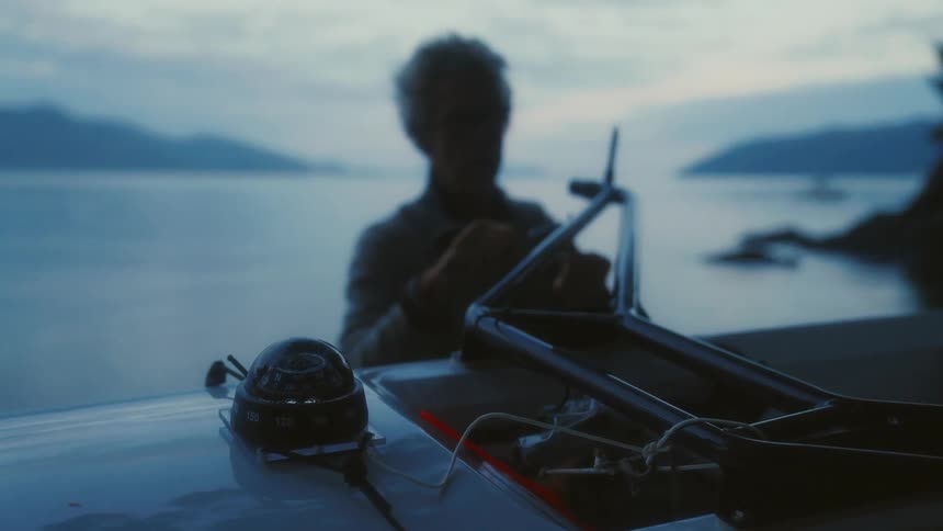 An elderly man threads a fishing line at a table next to a large body of water