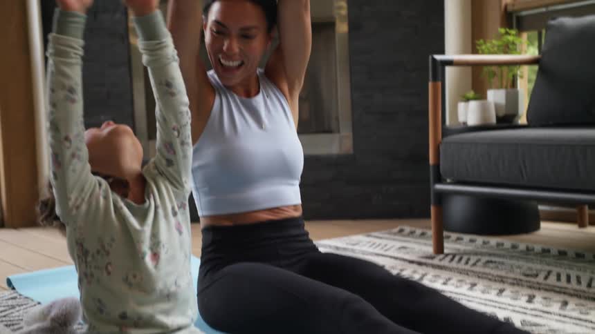 A woman tickles her daughter while they stretch on the floor of their living room