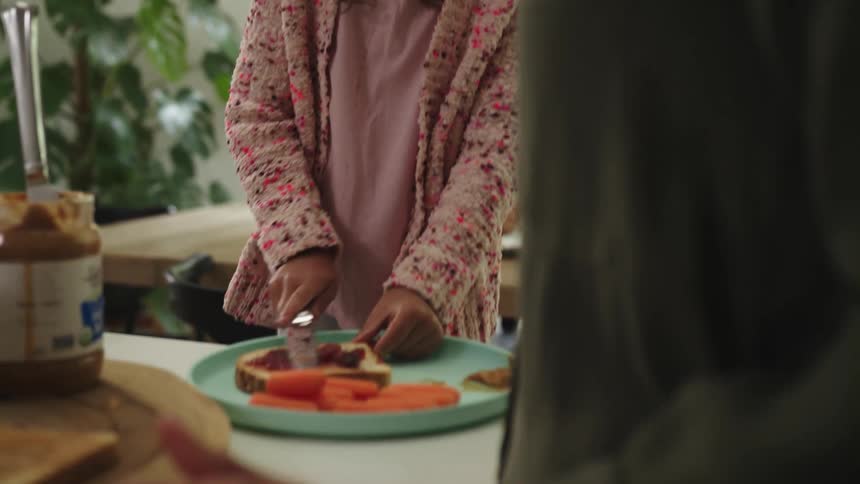 A young girl spreads jam on a piece of bread as her mother watches