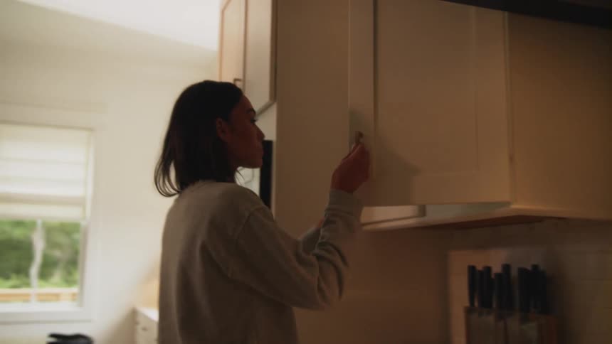 A woman walking up to a kitchen cupboard and removing a cup
