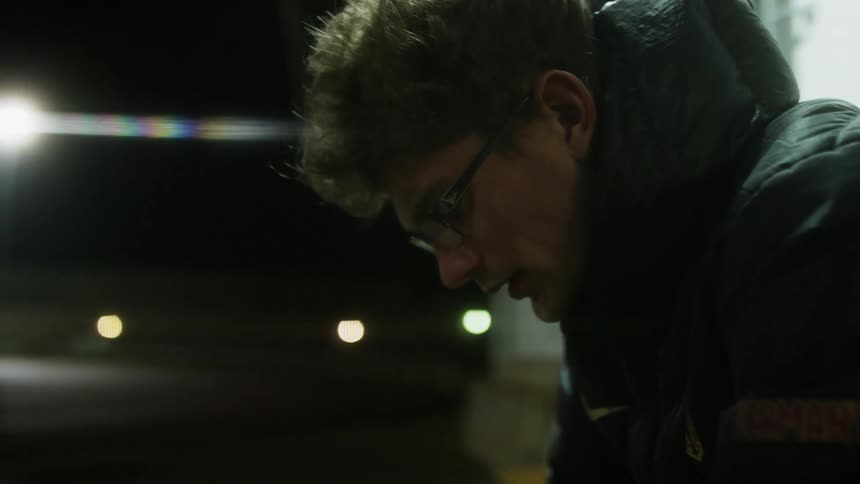 A man sitting outside at night wearing a coat and looking down