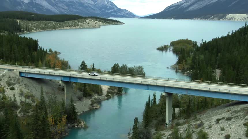 An automobile crosses a bridge over a forested lake with mountains in the distance