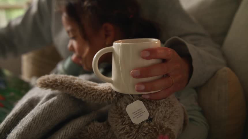 Mother wearing gray sweater hugs daughter holding a white cup of tea sitting on couch