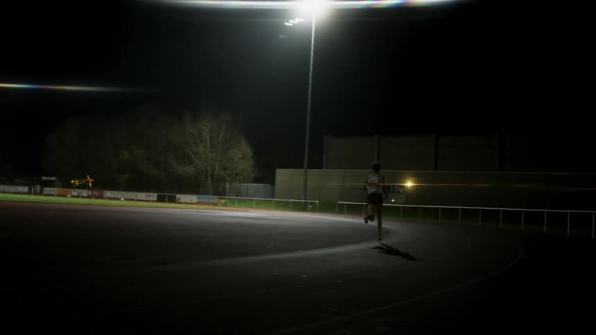 A track runner is running on the field at night as a light shines brightly from above