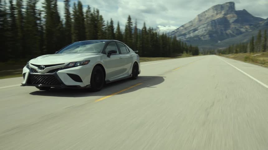 White sports car drives on highway next to the forest with mountain in the background