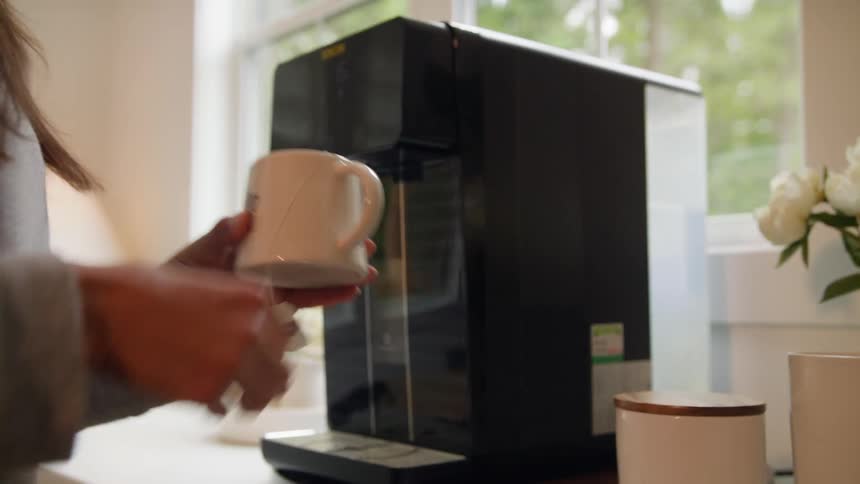 A woman places a tea bag in a mug then pushes a button to add hot water