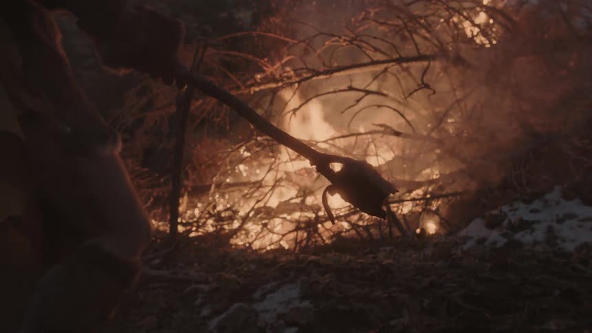 A man holding a smoking torch approaches a pile of burning brush