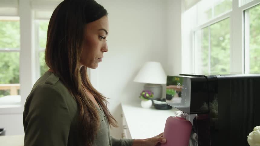 Woman with brown straight hair and green shirt pours hot water into bottle from coffee machine in the kitchen