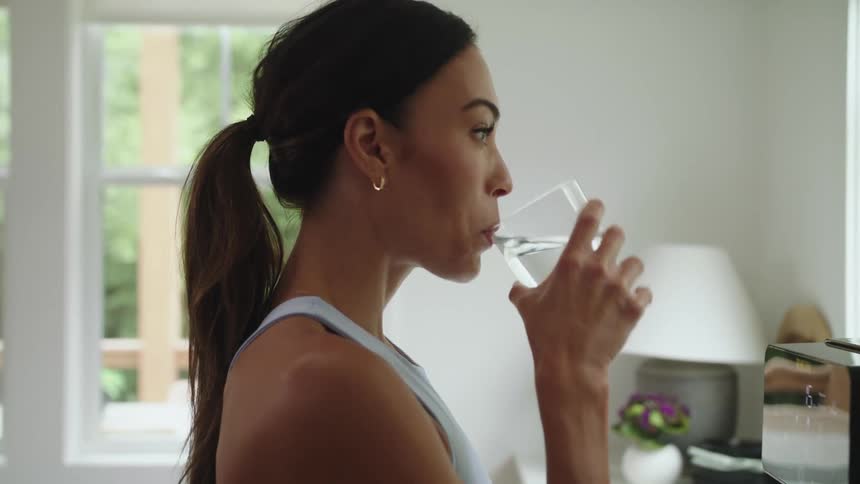 Woman with ponytail and sports bra drinks a glass of water in the kitchen at home