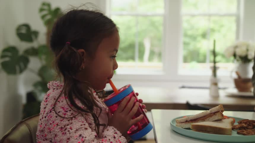 A young girl drinks from a straw cup while sitting at a dining table in a home kitchen