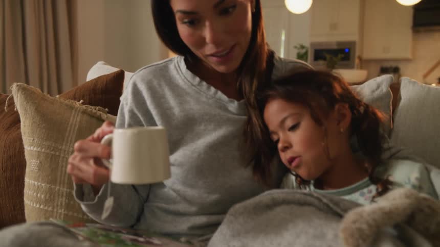 Mother holding a cup of tea talks to her daughter while reading a storybook on the couch at home