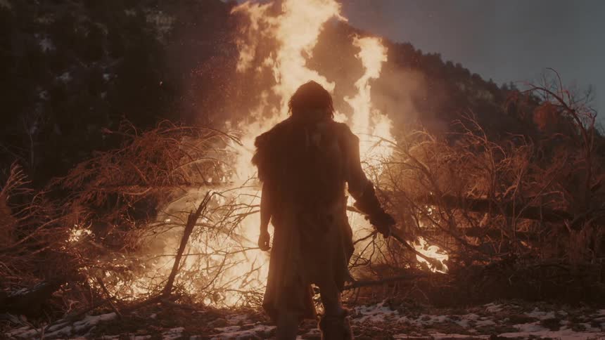 A man carrying a torch stands before a large pile of tree branches that is burning