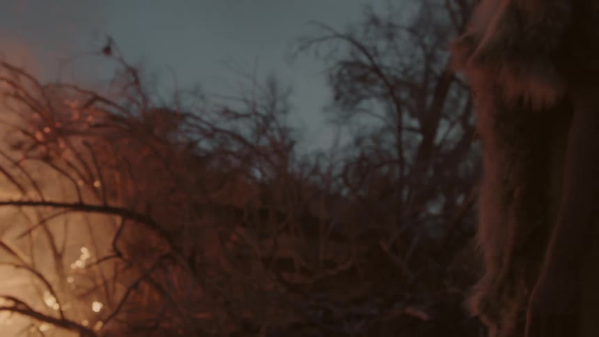 Man wearing furs stands by a burning pile of brush