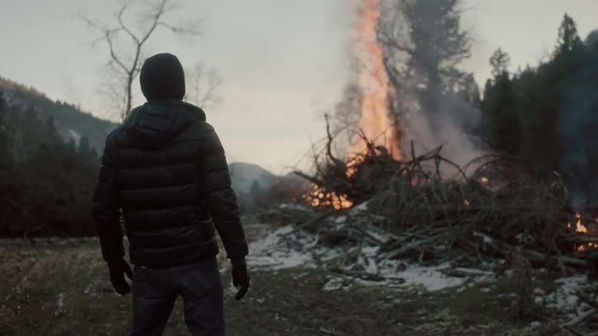 A man in a winter jacket stands looking at a burning pile of tree branches