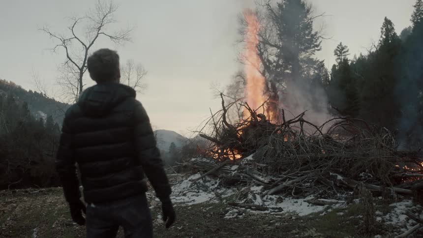 Man stands beside large brush pile as flames climb higher