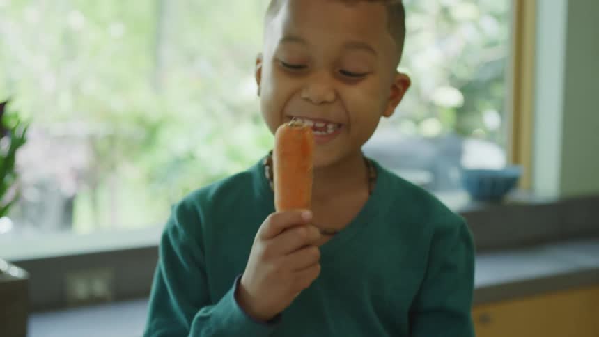 A boy pulls a carrot out of a box of produce and pretends to eat it