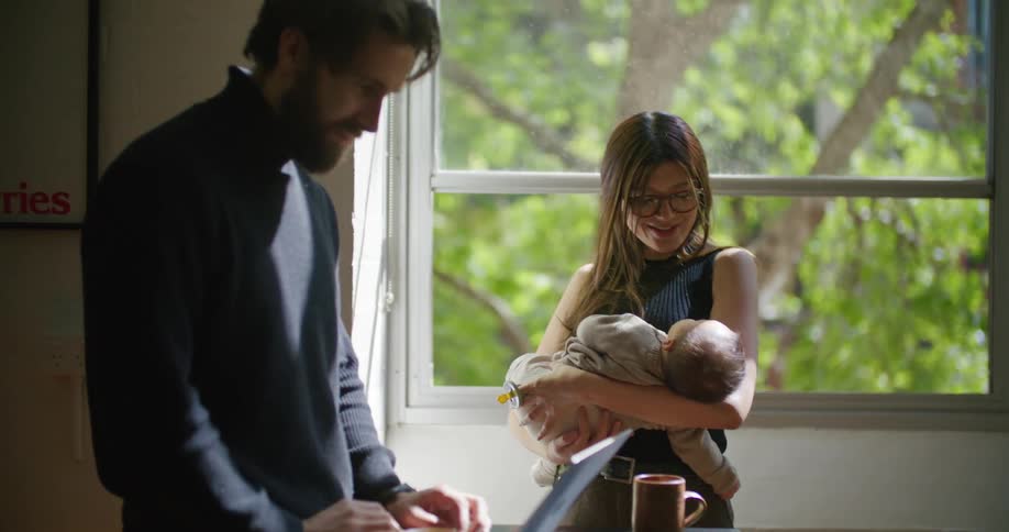 Young bearded man searching on a laptop next to her wife holding their newborn baby at home