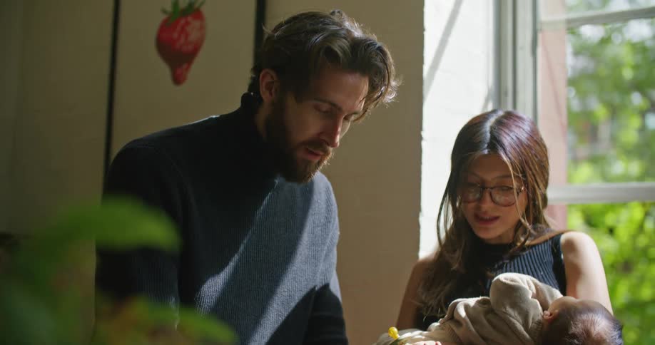 Father with beard and dark blue turtleneck sweater works on laptop as mom with brown hair glasses and green top holds baby in her arms in the kitchen at home