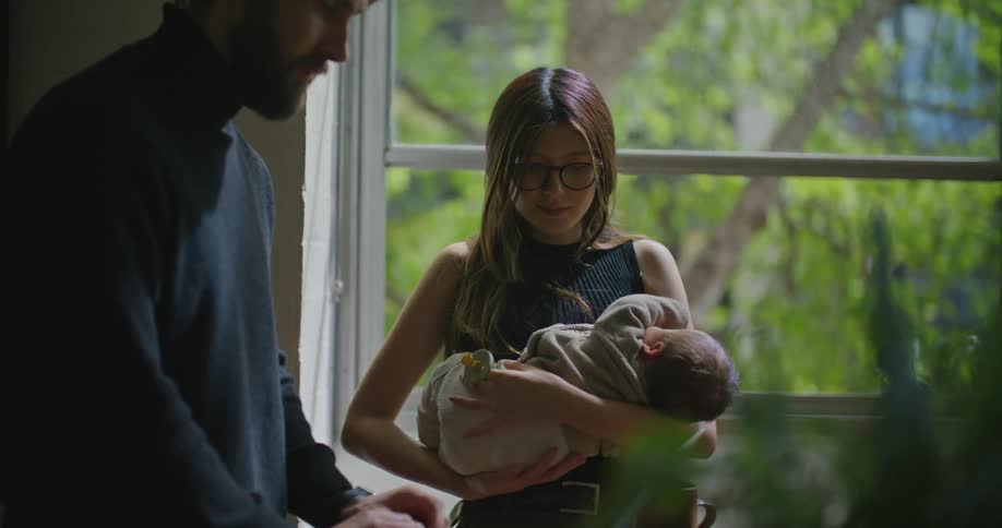 Father with beard and dark blue turtleneck sweater works on laptop as mom with brown hair glasses and green top holds baby in her arms in the kitchen at home