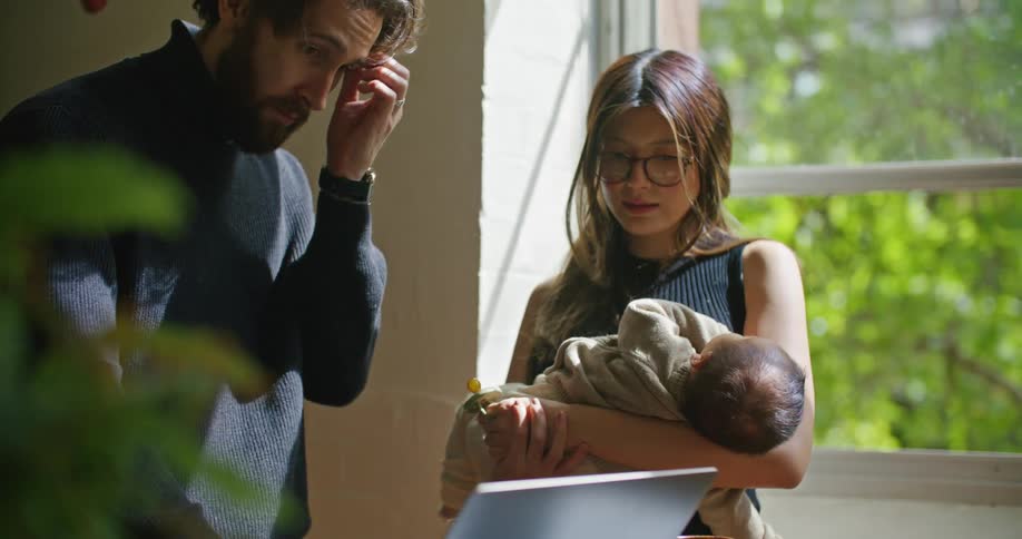 Young bearded man searching on a laptop next to her wife holding their newborn baby at home