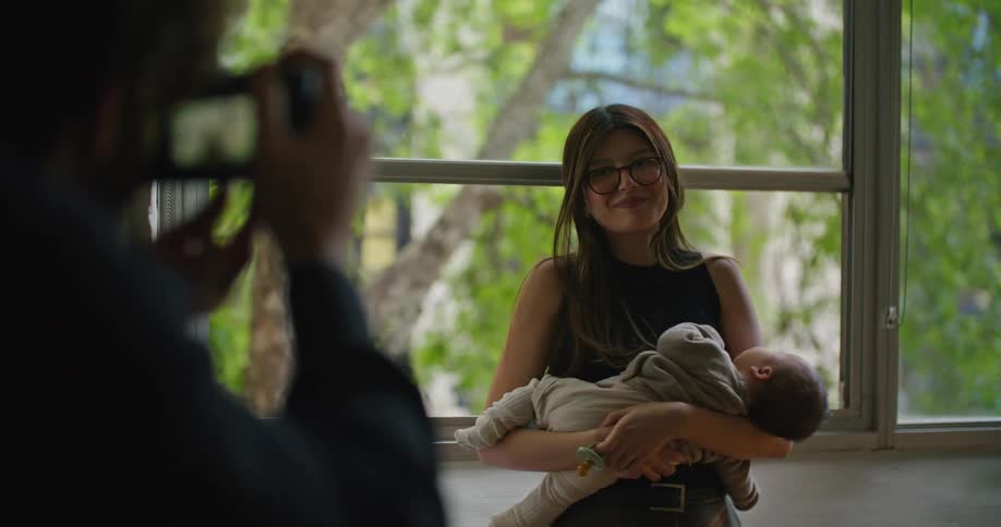 A woman standing in front of windows holding her baby looking up smiling as dad takes a picture