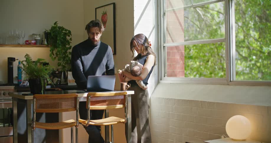 Father with beard and dark blue turtleneck sweater works on laptop as mom with brown hair glasses and green top holds baby in her arms in the kitchen at home