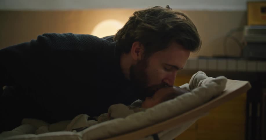 Father rocks child in baby rocker and leans over to kiss baby on forehead