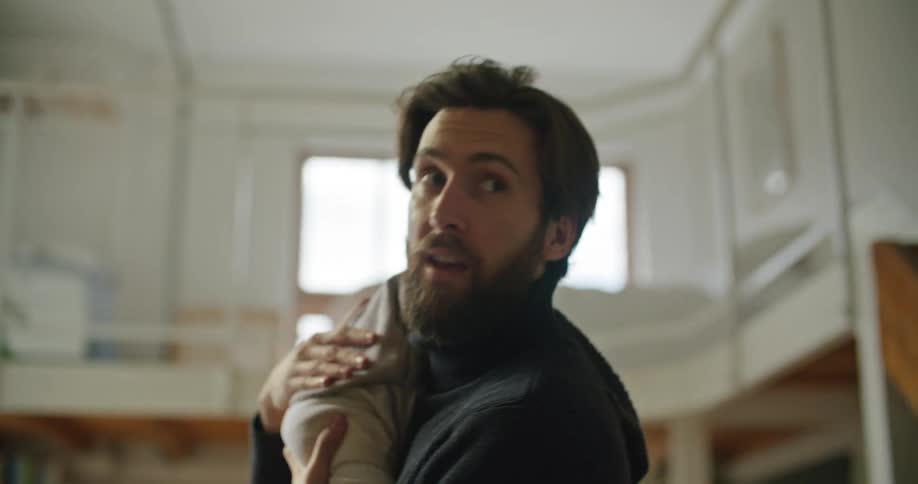 Young bearded man holding a baby over his shoulder turns around to talk to his wife at home