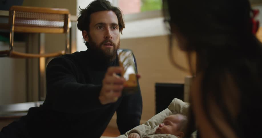 Man shows a picture on a smartphone to his wife while sitting near their baby at home