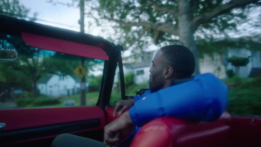 A man is sitting in the passenger side of a red convertible car as it travels through a neighborhood